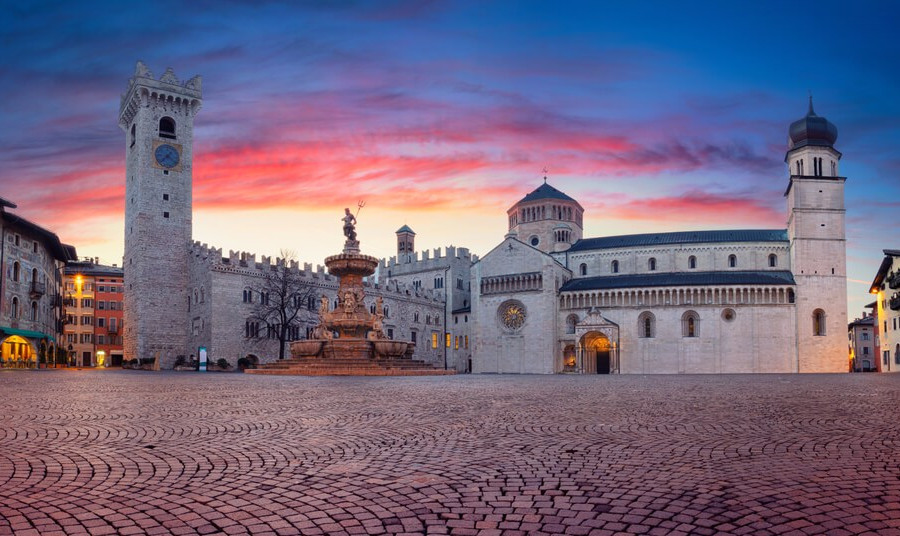 イタリア北部トレントのドゥオーモ広場。夕暮れの空の下、中世の建築と大聖堂が並ぶ歴史的な街並み